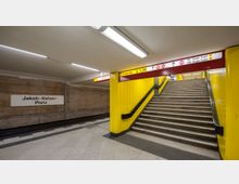 U-Bahn-Station mit gelb gestrichenen Wänden und einem Treppenaufgang, der zu einem Ausgang führt. Ein Schild mit der Aufschrift „Jakob-Kaiser-Platz“ ist an der Wand angebracht, und Deckenschilder zeigen Richtungen zu Bussen und dem Flughafen Tegel an.