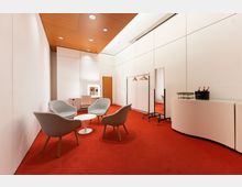 Umkleideraum mit rotem Teppichboden, weißen Wänden und holzverkleideter Decke. Der Raum enthält graue Sessel um einen kleinen weißen Tisch, einen Frisiertisch mit beleuchtetem Spiegel, eine Garderobe, einen Ganzkörperspiegel und eine weiße Anrichte mit Gläsern und Getränken.