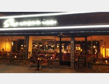 Beleuchtete Außenansicht eines Restaurants mit großen Glasfronten, die den Blick ins Innere freigeben. Drinnen sind Tische, Stühle und eine Bar mit hängenden Lampen sichtbar; vor der Tür stehen zwei kleine Tische und ein Aufsteller mit Menü.