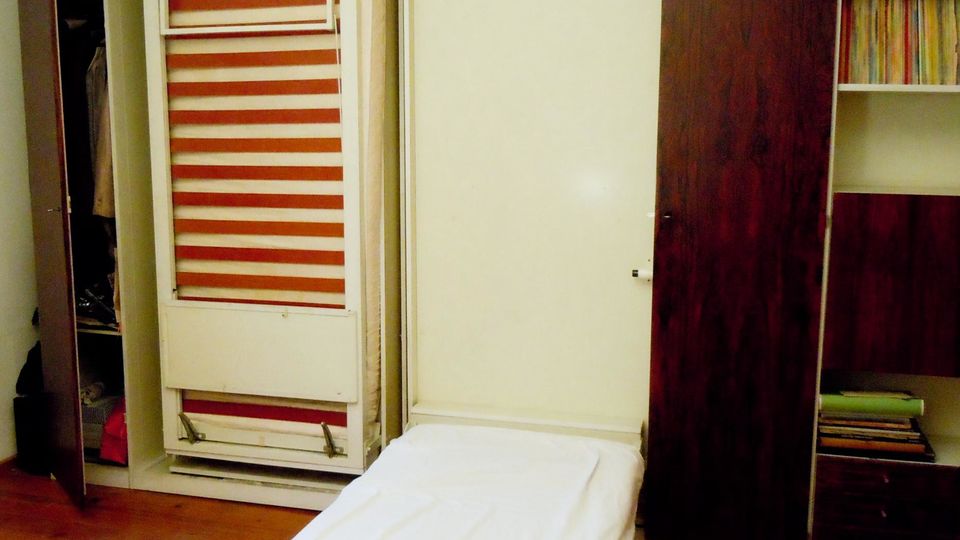 Innenraum eines Schlafzimmers mit ausgeklapptem Schrankbett im Vordergrund und großen Einbauschränken an der Wand. Links ist ein offener Schrank mit sichtbarem Lattenrost-Mechanismus, rechts ein Schrank mit Regalfächern und Holzfronten auf Holzboden.