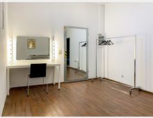 Ein schlicht eingerichteter Umkleideraum mit einem Schminktisch, beleuchtetem Spiegel, einem schwarzen Stuhl, einem großen Standspiegel und einer Kleiderstange mit Kleiderbügeln. Der Raum hat weiße Wände und einen dunklen Holzboden.