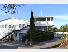 Weiße, moderne Gebäudeanlage mit Flachdach, bestehend aus einem Hauptgebäude und einer verglasten, erhöhten Plattform auf Stelzen. Im Vordergrund befinden sich eine Außentreppe, niedrige Büsche, eine schlanke Zypresse und ein gelbes Geländer, umgeben von Gehwegen und einer offenen, grünen Umgebung.