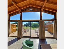 Innenraum mit Holzbalkendecke und großen bodentiefen Fenstern, die auf eine Terrasse mit Tischen und Stühlen führen. Im Hintergrund eine grüne Wiese mit Bäumen unter klarem, sonnigem Himmel.