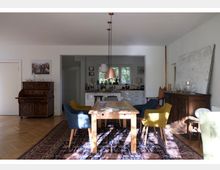 Esszimmer mit Holzfußboden, einem Holztisch und gemischten Polsterstühlen auf einem orientalischen Teppich. Im Hintergrund befindet sich eine offene Küche mit weißen Schränken und einem Fenster, das Tageslicht hereinlässt.