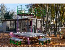Spielplatzbereich mit Betonpicknicktischen in Pink und Grün sowie einer Holzkonstruktion mit Aussichtsplattform und Wendeltreppe im Hintergrund. Herbstlaub bedeckt den Boden, umgeben von kahlen Bäumen und einem Gebäude mit großen Glasfronten.