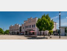 Historisches Bahnhofsgebäude mit heller Fassade und einer großen Uhr über dem Eingang, flankiert von Bäumen und Laternen. Vor dem Gebäude befinden sich Sitzbänke, Sonnenschirme und ein Platz mit Gehwegplatten, umgeben von einer klaren, blauen Himmelkulisse.