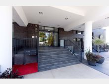 Eingangsbereich eines modernen Gebäudes mit dunklen Wandpaneelen, großen Glasfenstern und einer Treppe mit schwarzen Geländern. Im Vordergrund befinden sich ein roter Teppich, mehrere Pflanzen in Töpfen und Briefkästen.