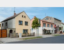 Straßenszene mit Wohn- und Geschäftshäusern; links ein beige verputztes Wohnhaus mit rotem Holztor und Gartenzaun, rechts ein rosafarbenes Gebäude mit Geschäft im Erdgeschoss samt Markise. Im Vordergrund Gehweg, Baum und Straße, im Hintergrund blauer Himmel.