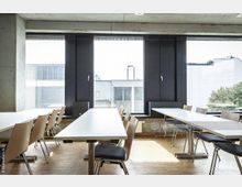 Heller Besprechungsraum mit langen weißen Tischen und hellen Holzstühlen, großen Fenstern mit Blick auf ein Gebäude und Bäume im Hintergrund, sowie sichtbarem Beton und Holzfußboden.