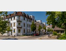 Straßenszene mit mehrstöckigen Wohngebäuden in heller Fassade, teils mit Balkonen und roten Dachziegeln. Vor den Häusern stehen Bäume, Straßenlaternen und Verkehrsschilder, im Vordergrund sind parkende Autos und Fahrräder sichtbar.