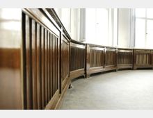 Holzvertäfelung mit vertikalen Lamellen in einem hell beleuchteten Raum, der durch große Fenster mit hohen Rahmen charakterisiert ist; der Boden ist mit Teppich bedeckt.