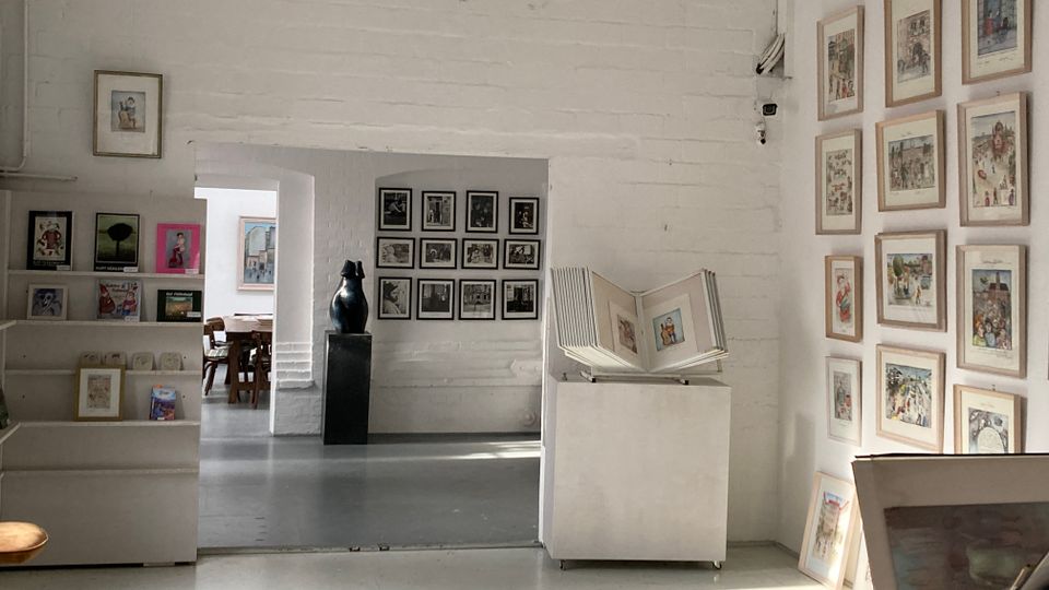 Innenraum einer hellen Galerie mit weißen Backsteinwänden und grauem Boden, an denen zahlreiche gerahmte Illustrationen hängen. Im Vordergrund stehen ein Regal mit Büchern, ein Sockel mit Skulptur und ein Podest mit aufgeschlagenem großformatigem Buch; durch einen Durchgang ist ein weiterer Raum mit Tischen und Bildern zu sehen.