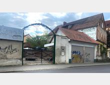 Einfahrt in einen Hinterhof mit einem schwarzen Gittertor, umgeben von Gebäuden mit roten Ziegeldächern und teils verwitterten Fassaden. Ein Werbeschild an der Hauswand weist auf ein Teppichzentrum hin, und die Umgebung enthält Graffiti sowie einen gepflasterten Gehweg.