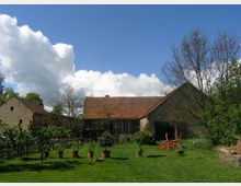 Alter Bauernhof mit Ziegeldächern und Fachwerkelementen, umgeben von einem großen Garten mit Rasen, Topfpflanzen und einigen Bäumen. Im Hintergrund blauer Himmel mit weißen Wolken.