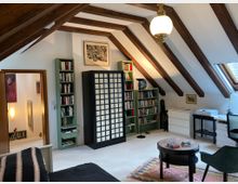 Dachzimmer mit sichtbaren Holzbalken, weißen Wänden und Dachfenster. Der Raum ist eingerichtet mit Bücherregalen, einem schwarzen Schrank mit weißen Paneelen, einer Sitzgruppe mit rundem Tisch und Teppich, sowie einem Schreibtisch mit Büromaterial.