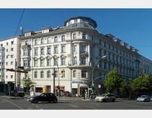 Eckgebäude eines historischen Wohn- und Geschäftshauses mit weißer Fassade, reich verzierten Fenstern und einem abgerundeten Dachgeschoss mit Kuppel; im Erdgeschoss befinden sich ein Bäckerei-Café und weitere Geschäfte. Im Vordergrund eine Straßenkreuzung mit vorbeifahrenden Autos und einer Straßenecke mit Bäumen.