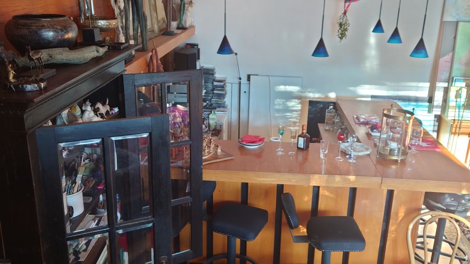 Innenraum eines gemütlich eingerichteten Wohn- oder Aufenthaltsbereichs mit Holzverkleidung, einer Bar mit hohen schwarzen Hockern und Pendelleuchten. Im Vordergrund ist ein Schrank mit Glastüren zu sehen, der mit Dekoobjekten und Büchern gefüllt ist.