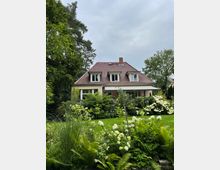 Zweistöckiges Einfamilienhaus mit roten Dachziegeln und mehreren Fenstern, umgeben von einem üppigen Garten mit grünen Büschen, Farnen und blühenden weißen Hortensien. Im Hintergrund befinden sich hohe Bäume, und der Himmel ist wolkig.