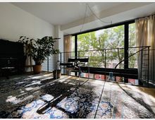 Helles Wohnzimmer mit großen bodentiefen Fenstern, durch die Bäume und Gebäudefassaden sichtbar sind. Im Raum stehen ein schwarzes Klavier, ein großer Topfpflanze, ein schwarzer Stuhl aus Metall und ein gemusterter Teppich auf dem Holzboden.