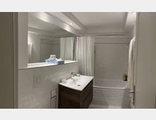 Modernes Badezimmer mit weißen Fliesen im U-Bahn-Stil, einer Badewanne mit Duschvorhang, einem Spiegel über einem Waschbecken auf einem Holzunterschrank und einem Handtuchhalter an der Wand. Die Beleuchtung erfolgt durch eingelassene Deckenstrahler.