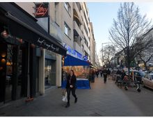 Straßenszene in einer städtischen Einkaufsstraße mit Geschäften und Restaurants, darunter ein "Burger Grill"-Lokal mit schwarzem Vordach. Im Vordergrund Gehweg mit Passanten, rechts parkende Autos und Bäume, im Hintergrund weitere Gebäude und Geschäfte.