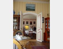 Ein historisch eingerichteter Raum mit Holzboden, klassischen Möbeln und Dekorationen. Geöffnete Flügeltüren führen in einen angrenzenden Raum mit gedecktem Esstisch, Gemälden an den Wänden und einer kleinen Gruppe von Personen.