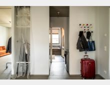 Flur in einer modernen Wohnung mit hellen Wänden, links ein kleiner Stauraum mit einem Vorhang, rechts eine Garderobe mit Jacken und Taschen, sowie einem roten Koffer auf dem Boden. Durch den Flur führt eine Tür zu einem Badezimmer mit einem Waschbecken, einer Waschmaschine und einem Fenster mit Jalousien.