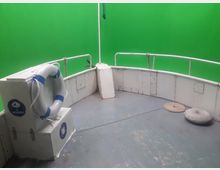 Der Vorderbereich eines kleinen Schiffdecks mit weißen Relinggeländern, einem Rettungsring an einer Kiste sowie einem grünen Hintergrund. Der Boden ist grau und zeigt einige Seilknoten und einen runden Gewichtsstern.