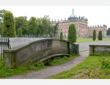Historisches Gebäude mit rosa Fassade und grüner Kuppel im Hintergrund, umgeben von parkähnlichen Gärten mit gepflegten Bäumen und Wegen. Im Vordergrund steht eine kleine steinerne Brücke über einen schmalen Graben.