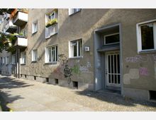 Wohnhaus in einer Stadt mit grauer Fassade, mehreren Fenstern und Balkonen mit Blumenkästen; die Wand ist mit farbigen Graffitis besprüht. Der Eingangsbereich des Gebäudes besteht aus einer weißen Gittertür, und der Bürgersteig davor ist mit Kopfsteinpflaster bedeckt.