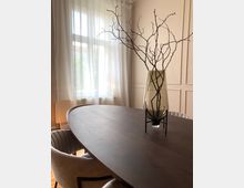 Ein elegant eingerichteter Raum mit einem ovalen Holztisch, umgeben von gepolsterten Stühlen. Auf dem Tisch steht eine hohe, durchsichtige Vase mit dekorativen Zweigen, im Hintergrund ein großes Fenster mit weißen, lichtdurchlässigen Vorhängen und einer cremefarbenen Wand.