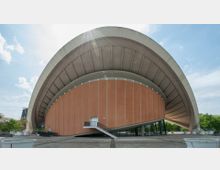 Außenansicht eines modernen Gebäudes mit einer geschwungenen, muschelartigen Dachkonstruktion und einer orangefarbenen, vertikal gegliederten Fassade. Im Vordergrund befinden sich eine Freitreppe, eine gepflasterte Fläche und mehrere Pflanzkübel; im Hintergrund sind Bäume und ein heller Himmel sichtbar.