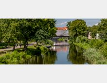 Ein Fluss mit ruhigem Wasser, umgeben von Bäumen und grüner Vegetation, führt zu einem weißen Gebäude mit rotem Dach im Hintergrund. Im Vordergrund sind Spazierwege und Sitzbänke in einem parkähnlichen Bereich sichtbar.