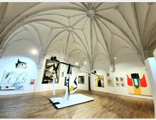 Raum einer Kunstgalerie mit gotischem Gewölbe, modernen abstrakten Gemälden an den Wänden und einer skulpturalen Installation in der Mitte auf einem Holzfußboden. Die Architektur umfasst Spitzbögen und weiße Wände, die die ausgestellten Werke hervorheben.