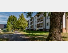 Wohngebäude mit drei Etagen und Balkonen, umgeben von Bäumen und Wiese; ein Weg führt entlang des Gebäudes, während Wäscheleinen mit aufgehängter Kleidung im Vordergrund sichtbar sind. Ein großer Baum steht rechts, und es ist eine ruhige, grüne Wohnumgebung.