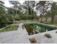 Ein moderner Garten mit einem rechteckigen Naturteich, umgeben von grauen Steinplatten. Im Hintergrund befinden sich Bäume, Sträucher und eine Rasenfläche sowie Holzbänke vor einer Holz- und Rankwand.