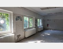Ein leerer, heller Raum mit gefliesten Wänden und einem gefliesten Boden. Große Fenster mit Blick ins Grüne lassen Tageslicht herein, darunter befinden sich weiße Heizkörper; an der Wand hängt ein Feuerlöscher.