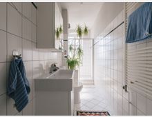 Schlichtes Badezimmer mit weißen Fliesen an Wänden und Boden, einem rechteckigen Waschbecken mit Spiegelschrank darüber, einem Handtuchhalter mit blauem Handtuch und einer Dusche im Hintergrund. Pflanzen in Töpfen und ein bunter Teppich verleihen dem Raum dekorative Akzente.