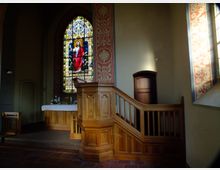 Innenraum einer Kirche mit einem hölzernen Kanzelaufbau und einem verzierten Buntglasfenster, das Jesus in rotem Gewand darstellt. Im Hintergrund befindet sich ein Altar mit weißen Blumen, flankiert von bemalten Wänden mit floralen Mustern.