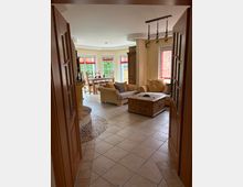 Wohnzimmer mit hellen Fliesenboden, einer beigen Eckcouch, einem Holztisch mit Schubladen und einem Essbereich mit Holzmöbeln im Hintergrund. Große Fenster mit roten Rollos lassen viel Tageslicht herein.