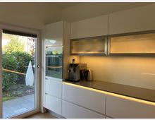 Moderne Küche mit weißen Hochglanzschränken und eingebautem Backofen. Eine Glastür führt zu einem Garten mit dichtem Grün, während warmes Unterbau-Licht die Arbeitsflächen und Küchengeräte beleuchtet.