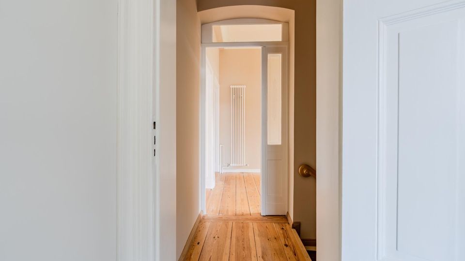 Schmaler Innenflur in einer Wohnung mit hellem Holzdielenboden, hohen weißen Türrahmen und einem Rundbogen am Ende des Gangs. Durch eine offene Tür ist ein angrenzender Raum mit hohem, schmalem Heizkörper an der Wand zu sehen.