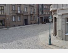 Straßenszene mit Kopfsteinpflaster und mehreren historischen Wohn- und Geschäftshäusern mit verzierten Fassaden. An der Straße stehen grüne Laternen im Stil des 19. Jahrhunderts.