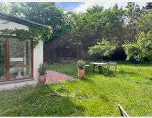 Garten mit Rasenfläche und dichtem Baumbestand, links ein kleines Haus mit großen Fenstern und mit Efeu bewachsener Fassade. In der Mitte stehen ein Tisch mit Klappstühlen und eine Bank, daneben ein gepflasterter Weg und mehrere Pflanzkübel.