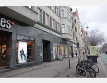 Straßenszene mit mehrstöckigen Wohn- und Geschäftsgebäuden; im Vordergrund ein Laden mit großen Schaufenstern und grauer Fassade, daneben Fahrräder an einem Fahrradständer. Im Hintergrund Gehweg, Straßenbeleuchtung und kahle Bäume entlang der Straße.