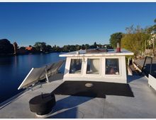 Ein Boot mit weißen Aufbauten und Fenstern liegt an einem ruhigen Flussufer. Im Hintergrund sind ein rotes Gebäude, Bäume und eine klare, blaue Himmelsszene sichtbar.