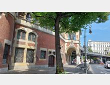 Backsteingebäude im historischen Stil mit gotischem Eingangsportal und Spitzbogenfenstern, flankiert von einem Baum im Vordergrund. Rechts im Bild eine erhöhte Metallbrücke und klassizistische Stadtarchitektur im Hintergrund, daneben parkende Fahrräder und Autos an einer Straßenkreuzung.