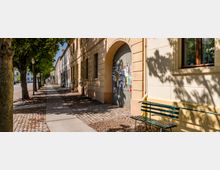 Gepflasterter Gehweg entlang einer von Bäumen gesäumten Straße mit historischen, hell verputzten Fassaden. Im Vordergrund stehen eine grüne Sitzbank und ein großes Torbogenportal mit angeschlagenen Plakaten.