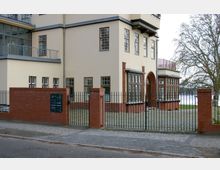 Eckansicht eines Gebäudes mit hellbeiger Fassade, großen Fenstern und Backsteinelementen, einschließlich eines Rundbogeneingangs. Vor dem Gebäude befindet sich ein aus roten Ziegeln und Metallzaun gestaltetes Tor, dahinter ein gepflasterter Hof mit Blick auf einen See und Bäume im Hintergrund.