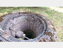 Ein alter, steinerner Schacht mit runder Öffnung, überwuchert von Moos und kleinen Pflanzen. Im Inneren sind Metalltritte einer Leiter sichtbar, die nach unten führen, und am Rand des Schachtes befindet sich eine Metallstange.